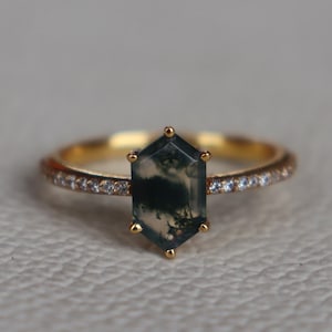 Può includere: Un anello d'oro con una pietra di agata muschiata esagonale. La fascia è decorata con piccole pietre preziose trasparenti. L'agata muschiata presenta un motivo verde e bianco. L'anello è posto su uno sfondo neutro.