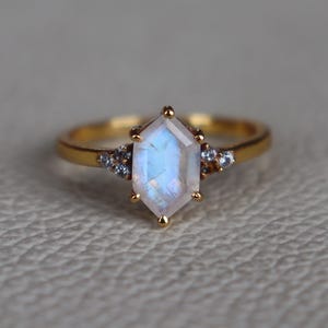 Op de afbeelding: Een gouden ring met een zeshoekige, iriserende maansteen in het midden. Kleine accentstenen flankeren de hoofdsteen aan weerszijden. De ring is geplaatst tegen een neutrale achtergrond, waardoor het ontwerp van het sieraad wordt benadrukt.