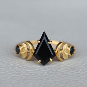 May include: A gold ring featuring a black gemstone at its centre, flanked by two crescent moon accents. The ring is adorned with black stones on either side of the crescent moons.
