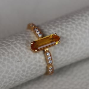 May include: A gold ring featuring a rectangular, amber-colored gemstone. The band is adorned with small, clear stones. The ring is set against a neutral background, highlighting its design and color.