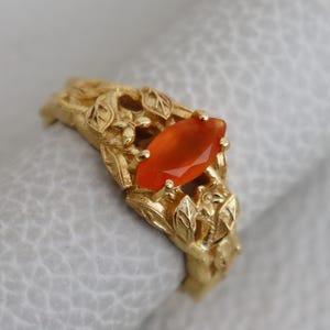 Könnte beinhalten: Ein goldener Ring mit einem geschnitzten Blattdesign und einem leuchtend orangefarbenen Edelstein in der Mitte.
