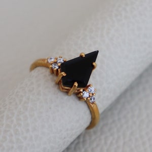 May include: A gold ring with a black gemstone in the shape of a diamond. The ring is set with smaller clear gemstones around the main stone.