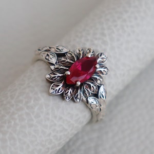 Puede incluir: Un anillo de plata con una piedra preciosa roja rodeada de un diseño de hojas.