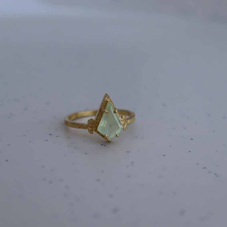 May include: A gold ring with a light green gemstone in a geometric shape.
