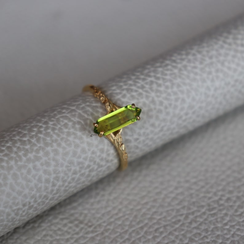 Pu&ograve; includere: Un anello d'oro con una pietra preziosa rettangolare verde chiaro. La fascia dell'anello presenta un design floreale ornato. La pietra preziosa &egrave; incastonata orizzontalmente, su uno sfondo neutro.