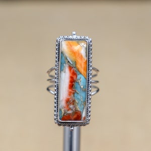 Pode incluir: Um anel de prata com uma pedra retangular turquesa, laranja e branca engastada em uma moldura de prata. O anel tem uma banda decorativa.