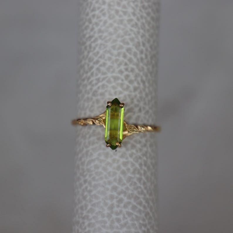 Pu&ograve; includere: Un anello d'oro con una pietra preziosa rettangolare verde chiaro. La fascia dell'anello presenta un delicato design a torsione con accenti di foglie. La gemma misura circa 1,3 cm di lunghezza.