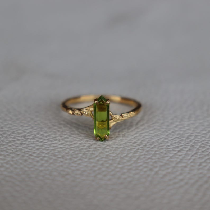 May include: A gold ring with a rectangular, light green gemstone. The band has a textured, vine-like pattern. The gemstone is set vertically in the centre of the ring, creating a simple yet elegant look. The ring is displayed on a neutral background.