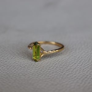 Pu&ograve; includere: Un anello d'oro con una brillante gemma rettangolare verde chiaro. L'anello ha un design a fascia intrecciata e un'incastonatura delicata. L'anello &egrave; esposto su una superficie neutra. La gemma misura circa 1 cm di lunghezza.
