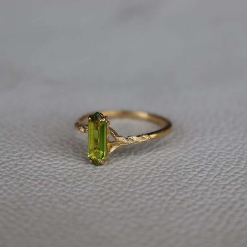 Pu&ograve; includere: Un anello d'oro con una brillante gemma rettangolare verde chiaro. L'anello ha un design a fascia intrecciata e un'incastonatura delicata. L'anello &egrave; esposto su una superficie neutra. La gemma misura circa 1 cm di lunghezza.