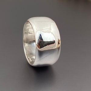 Könnte beinhalten: Ein hochglanzpolierter, breiter Silberring mit abgerundeter Oberseite. Der Ring reflektiert die Umgebung und erzeugt einen Spiegeleffekt. Das Design ist minimalistisch und für den täglichen Gebrauch geeignet. Die Breite beträgt ca. 1 cm.