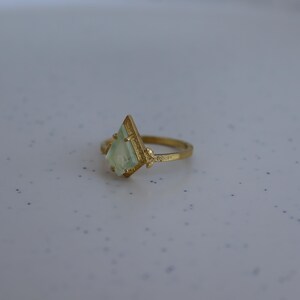 May include: A gold ring with a light green gemstone in a diamond shape. The ring band is decorated with a floral design.