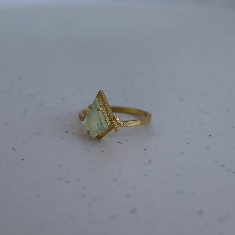 May include: A gold ring with a light green gemstone in a diamond shape. The ring band is decorated with a floral design.