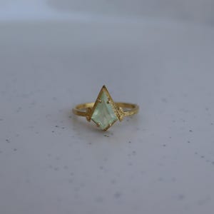 May include: A gold ring with a light green gemstone set in a pointed, geometric shape. The ring has a textured finish.