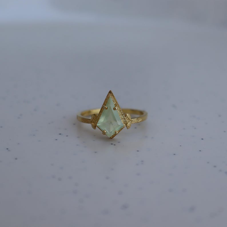 May include: A gold ring with a light green gemstone set in a pointed, geometric shape. The ring has a textured finish.