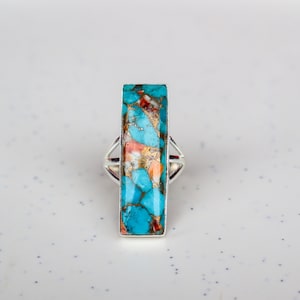 Może przedstawiać: Srebrny pierścionek z prostokątnym turkusem osadzonym w środku. Kamień ma unikalny wzór w kolorze niebieskim, pomarańczowym i białym.