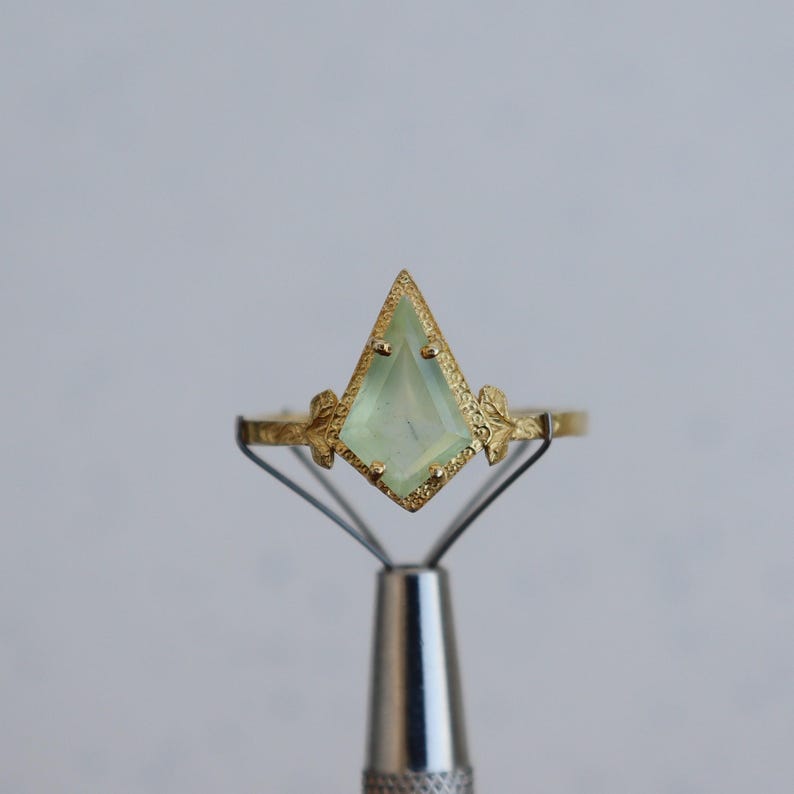 May include: A gold ring with a light green gemstone in a faceted, kite-shaped setting. The ring band is decorated with small, leaf-like details.