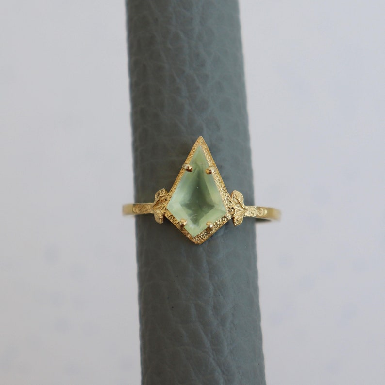 May include: A gold ring with a light green gemstone set in a diamond shape. The ring has a delicate floral design on the band.