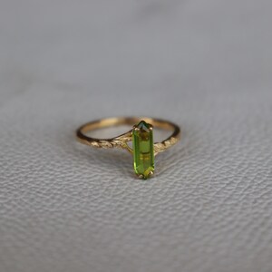 Pu&ograve; includere: Un anello d'oro con una vibrante pietra preziosa di peridoto verde. L'anello ha una fascia delicata e strutturata con un motivo floreale. Il peridoto &egrave; tagliato a forma rettangolare e incastonato verticalmente al centro dell'anello.