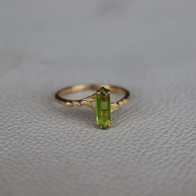 Pu&ograve; includere: Un anello d'oro con una vibrante pietra preziosa di peridoto verde. L'anello ha una fascia delicata e strutturata con un motivo floreale. Il peridoto &egrave; tagliato a forma rettangolare e incastonato verticalmente al centro dell'anello.