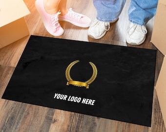 Puede incluir: Felpudo rectangular negro con un diseño de corona de laurel dorada y una pancarta dorada en blanco. El texto "YOUR LOGO HERE" está impreso en blanco. La alfombra está sobre un suelo de madera.