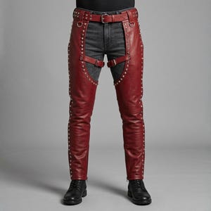 May include: Deep red leather chaps featuring silver-toned metal studs along the outer seams and thigh edges. The chaps include a buckled waist belt, two horizontal thigh straps with buckles, and D-ring hardware, worn over dark gray denim jeans.