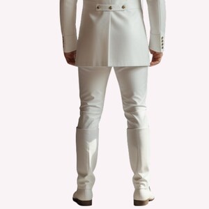 Puede incluir: Un traje blanco, que incluye una chaqueta con botones dorados, pantalones y botas. El conjunto es de color blanco brillante. Las botas altas hasta la rodilla tienen un peque&ntilde;o tac&oacute;n. El atuendo es formal.