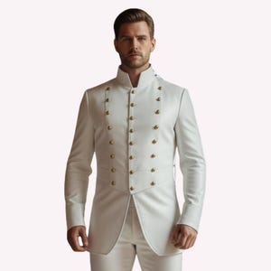 Puede incluir: Un traje blanco con cuello mao y dise&ntilde;o cruzado. La chaqueta presenta botones dorados y un corte a medida. El traje es de estilo formal, adecuado para ocasiones especiales.