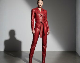 Op de afbeelding: Een rode leren outfit bestaande uit een jasje met een reverskraag en een bijpassende broek. De jas heeft een ritssluiting en een riem. De broek is hoog getailleerd en getailleerd. De outfit wordt gecompleteerd met rode leren laarzen.