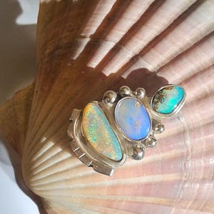 May include: A silver ring featuring three oval-shaped opals with iridescent flashes of green, blue, and orange. The setting includes small silver spheres accenting the stones, with a textured silver band base visible beneath the opal cluster.
