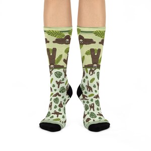 Könnte beinhalten: Ein Paar grüne und braune Socken mit einem Muster aus Faultieren, die Yoga-Posen einnehmen. Die Socken befinden sich auf einem weißen Hintergrund.