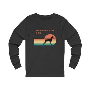 Op de afbeelding: Een donker grijs longsleeve t-shirt met een retro zonsondergang afbeelding en een silhouet van een Weimaraner hond. De tekst "Weimaraner Dad" is op het shirt gedrukt.