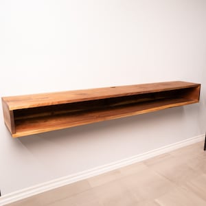 May include: A long, wooden floating shelf with a single open shelf in the middle. The shelf is mounted on a white wall and is made of light brown wood.