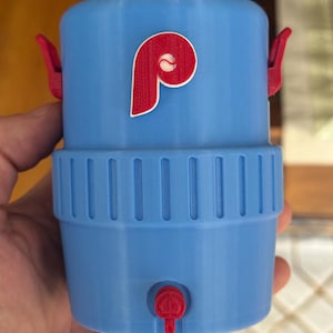 Può includere: Erogatore di bevande blu chiaro con logo Phillies rosso. L'erogatore ha un rubinetto rosso e due maniglie rosse. Il logo presenta una "P" rossa con una palla da baseball bianca.