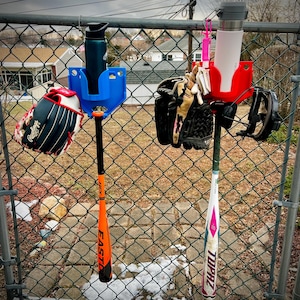 May include: A blue and red baseball equipment organiser hanging on a chain-link fence. The organiser holds a water bottle, baseball gloves, a baseball bat, and a baseball helmet. The baseball bat is orange and black, and the other bat is white and pink.