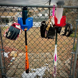 May include: Baseball equipment hanging on a chain-link fence. Includes a blue water bottle holder, a black and red baseball glove, an orange and black baseball bat, a red water bottle holder, a white and pink baseball bat, and baseball gloves.