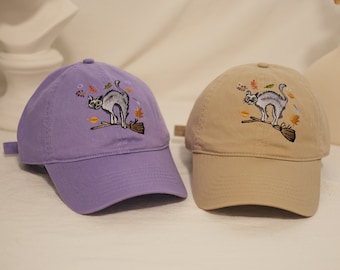 Op de afbeelding: Twee geborduurde baseballpetten met een zwart kattenontwerp. De ene pet is paars en de andere beige.