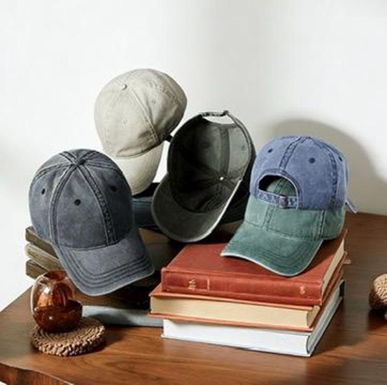 May include: A collection of baseball caps in various colors, including gray, navy, and a two-tone blue and green. The caps are arranged on a stack of books and a wooden table. The caps have a classic, worn look.