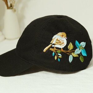 Könnte beinhalten: Eine schwarze Baseballkappe mit einem gestickten Design eines kleinen Vogels, der auf einem Zweig mit blauen und grünen Blumen sitzt.
