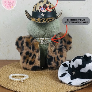 May include: A plush toy wearing a leopard print cowboy hat with a star, a matching jacket, and a pearl necklace with letter charms. A white and black cow print cowboy hat and a pearl bracelet are also present.