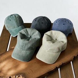 May include: Five assorted baseball caps in shades of green, blue, and gray are arranged on a wooden surface. The caps have a classic six-panel design with a curved brim and adjustable strap. The caps are made of a soft, worn fabric.