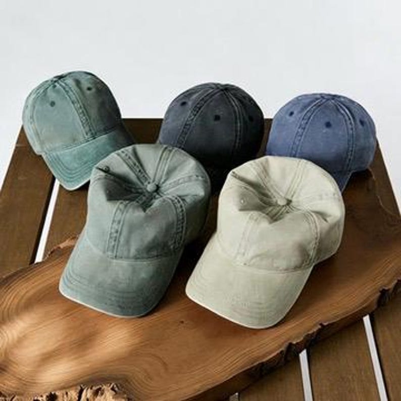 May include: Five assorted baseball caps in shades of green, blue, and gray are arranged on a wooden surface. The caps have a classic six-panel design with a curved brim and adjustable strap. The caps are made of a soft, worn fabric.