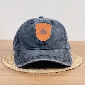 May include: A faded black baseball cap with a brown leather patch featuring a mountain design. The cap sits on a round, woven mat. The cap has a curved brim and adjustable strap.