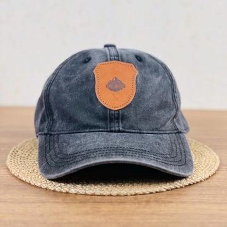May include: A faded black baseball cap with a brown leather patch featuring a mountain design. The cap sits on a round, woven mat. The cap has a curved brim and adjustable strap.
