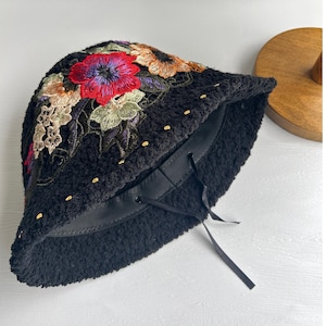 Puede incluir: Sombrero negro de cubo con un dise&ntilde;o de bordado floral. El sombrero tiene un lazo de cinta negro y detalles dorados.
