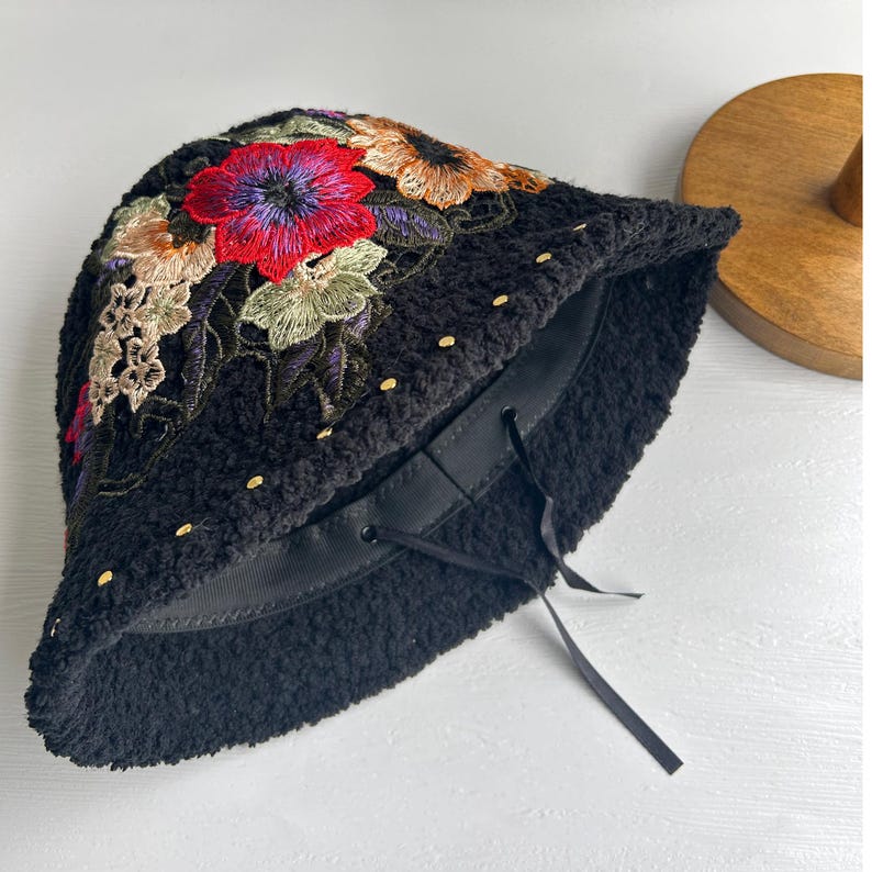 Puede incluir: Sombrero negro de cubo con un dise&ntilde;o de bordado floral. El sombrero tiene un lazo de cinta negro y detalles dorados.