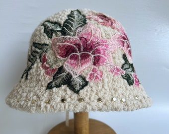 Puede incluir: Un gorro de cubo blanco y peludo con flores bordadas en rosa y verde. El gorro tiene detalles metálicos dorados alrededor del borde.