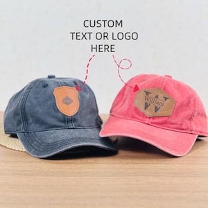 Puede incluir: Dos gorras de béisbol con parches de cuero. Una es azul marino, la otra es roja. La gorra azul marino tiene un parche en forma de escudo. La gorra roja tiene un parche rectangular con el nombre "William". El texto "CUSTOM TEXT OR LOGO HERE" está arriba.