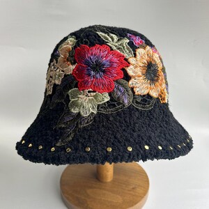 Puede incluir: Sombrero cubo negro con flores bordadas en tonos rojos, amarillos, verdes y morados. El sombrero tiene una textura peluda y tachuelas doradas alrededor del borde.