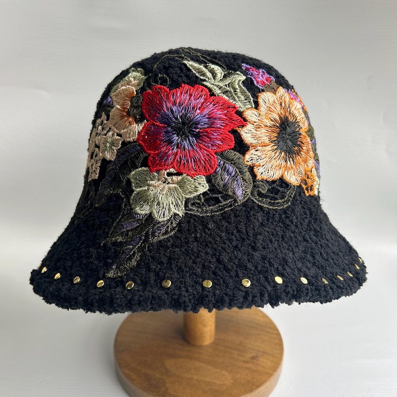 Puede incluir: Sombrero cubo negro con flores bordadas en tonos rojos, amarillos, verdes y morados. El sombrero tiene una textura peluda y tachuelas doradas alrededor del borde.